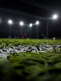 A close-up perspective of artificial grass with bright stadium lights illuminating from above. The focus is primarily on the green and white blades of the synthetic turf, with the background showing blurred outlines of light fixtures and possible figures.