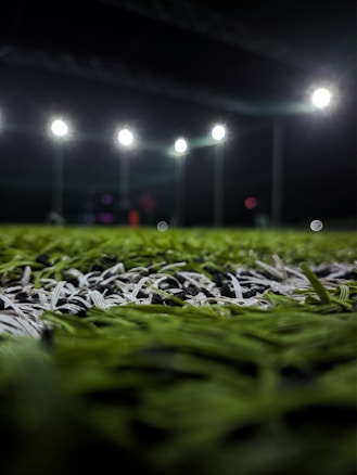 A close-up perspective of artificial grass with bright stadium lights illuminating from above. The focus is primarily on the green and white blades of the synthetic turf, with the background showing blurred outlines of light fixtures and possible figures.
