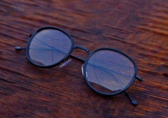 A pair of round black-rimmed eyeglasses rests on a wooden surface with visible texture, showcasing reflections on the lenses.