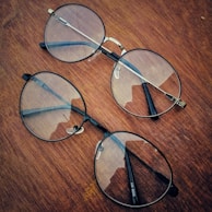 Two pairs of round eyeglasses with thin metal frames are placed on a wooden surface. The lenses have a slight reflection, and the wood has a rich, warm tone with visible grain patterns.