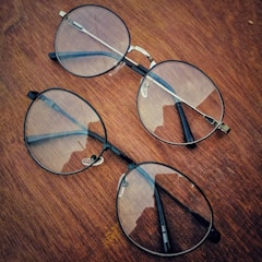 Two pairs of round eyeglasses with thin metal frames are placed on a wooden surface. The lenses have a slight reflection, and the wood has a rich, warm tone with visible grain patterns.