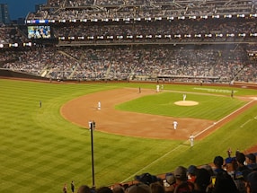 A baseball game is taking place in a large stadium filled with spectators. The field is well-lit, with players positioned at various points. The stands are packed with an enthusiastic crowd, and a large video screen displays the game.
