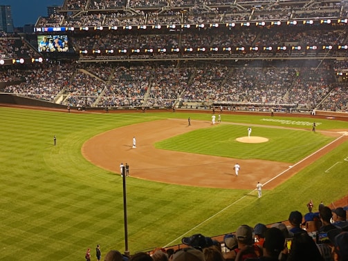 A baseball game is taking place in a large stadium filled with spectators. The field is well-lit, with players positioned at various points. The stands are packed with an enthusiastic crowd, and a large video screen displays the game.