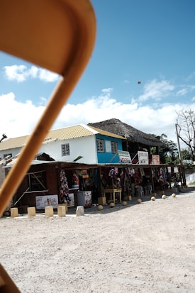 A street market scene with several shops displaying colorful fabrics and goods under a rustic thatched roof. A yellow chair is prominently in the foreground, partially obstructing the view. The sky above is blue with some scattered clouds, and there's a dirt road in front of the market.