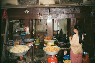 A woman with long black hair, wearing a patterned yellow top and pink pants, stands at a cluttered workstation frying food. In front of her are bowls filled with various food items, such as rice and fried snacks. The scene appears to be an inside view of a small, modest food stall or kitchen, with various kitchen utensils, pots, and ingredients scattered around. The surroundings are dimly lit with a single hanging light bulb, and there is an old, worn-out look to the interior decor.