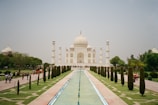 A grand white marble mausoleum stands majestically, flanked by four minarets. The structure is surrounded by lush gardens and a long reflecting pool that leads directly to it. Visitors stroll along the pathways, enjoying the serene and majestic atmosphere.