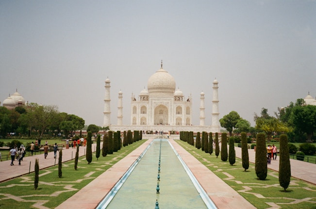 A grand white marble mausoleum stands majestically, flanked by four minarets. The structure is surrounded by lush gardens and a long reflecting pool that leads directly to it. Visitors stroll along the pathways, enjoying the serene and majestic atmosphere.