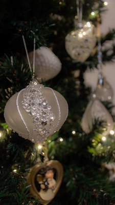 A collection of elegant white and silver Christmas ornaments are hanging on a lush green Christmas tree adorned with warm white lights. Each ornament glistens with intricate beading and patterns, adding a touch of sparkle and sophistication.