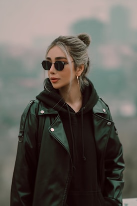 A person with dark sunglasses and hair tied up in a bun wears a black hoodie layered with a dark leather jacket. They stand against a blurred, neutral-toned urban background, giving a stylish and understated appearance.