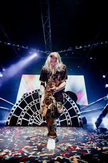 A musician with long hair is playing a saxophone on stage. The stage is decorated with a large, illuminated backdrop featuring abstract designs and bright lights. Colorful confetti is scattered across the floor, adding to the festive atmosphere. The musician is dressed in a dark shirt and patterned pants, and white sneakers.
