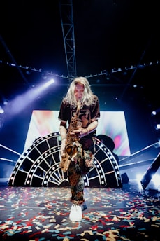 A musician with long hair is playing a saxophone on stage. The stage is decorated with a large, illuminated backdrop featuring abstract designs and bright lights. Colorful confetti is scattered across the floor, adding to the festive atmosphere. The musician is dressed in a dark shirt and patterned pants, and white sneakers.