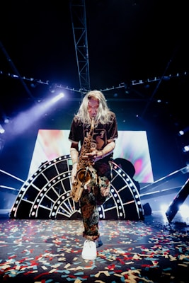 A musician with long hair is playing a saxophone on stage. The stage is decorated with a large, illuminated backdrop featuring abstract designs and bright lights. Colorful confetti is scattered across the floor, adding to the festive atmosphere. The musician is dressed in a dark shirt and patterned pants, and white sneakers.