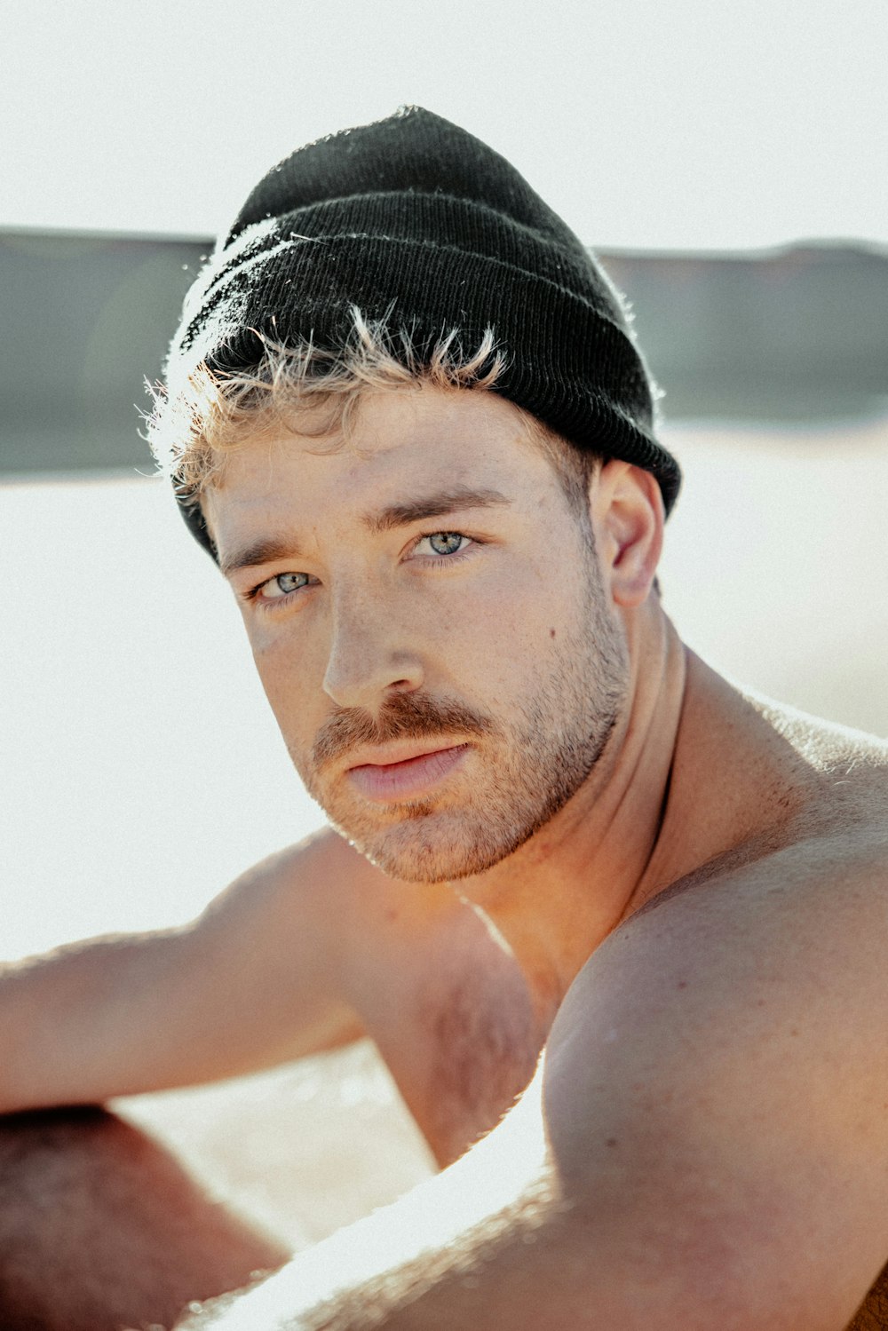 a shirtless man wearing a beanie sitting on a beach