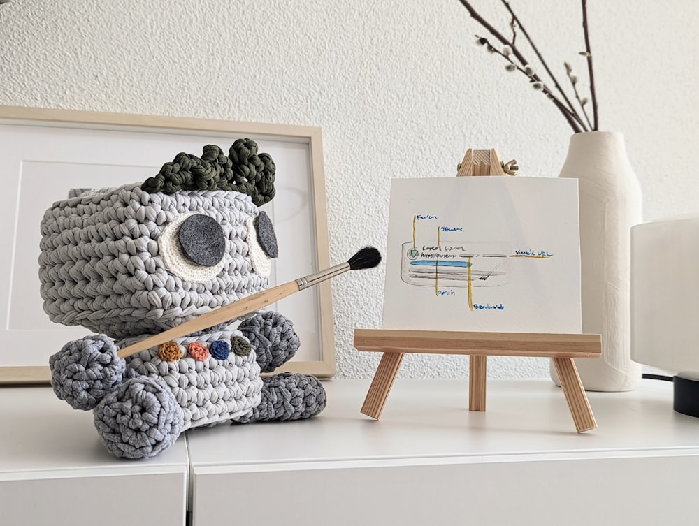 a crocheted koala holding a paintbrush next to a white easel