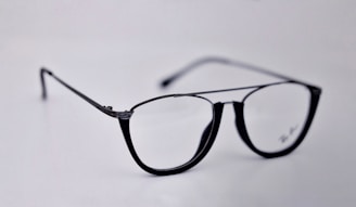 A pair of black-rimmed eyeglasses with a sleek and modern design. The glasses are placed on a plain light background, highlighting their stylish shape and glossy finish.