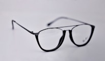 A pair of black-rimmed eyeglasses with a sleek and modern design. The glasses are placed on a plain light background, highlighting their stylish shape and glossy finish.
