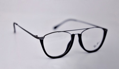 A pair of black-rimmed eyeglasses with a sleek and modern design. The glasses are placed on a plain light background, highlighting their stylish shape and glossy finish.