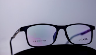 A pair of black-rimmed eyeglasses with the brand name Prada on the lens, set against a plain white background. The lenses display a subtle reflection with a hint of color, possibly from the surrounding lighting.