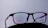 A pair of black-rimmed eyeglasses with the brand name Prada on the lens, set against a plain white background. The lenses display a subtle reflection with a hint of color, possibly from the surrounding lighting.