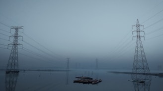 Several high-voltage power lines and steel towers stretch across a hazy and misty landscape. The reflection of the towers can be seen in a body of water below, creating a symmetrical and serene visual effect. The overall setting appears calm and slightly eerie due to the overcast sky and limited visibility.