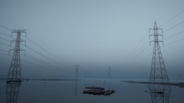 Several high-voltage power lines and steel towers stretch across a hazy and misty landscape. The reflection of the towers can be seen in a body of water below, creating a symmetrical and serene visual effect. The overall setting appears calm and slightly eerie due to the overcast sky and limited visibility.