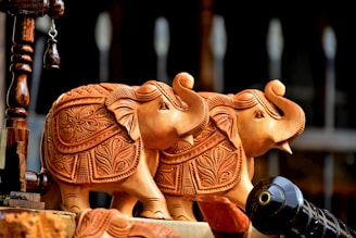 Intricately carved wooden elephant statuettes featuring detailed floral designs on their bodies. The figures are positioned close to each other, with a warm, earth-toned appearance.