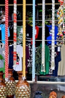 A shop display featuring several colorful garlands hanging in a row, with garments and patterns visible behind them. Large decorative vases with a pineapple motif are positioned at the bottom. Various types of clothing, including shirts, are hanging in the background, showcasing a variety of colors and designs.