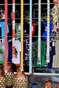 A shop display featuring several colorful garlands hanging in a row, with garments and patterns visible behind them. Large decorative vases with a pineapple motif are positioned at the bottom. Various types of clothing, including shirts, are hanging in the background, showcasing a variety of colors and designs.