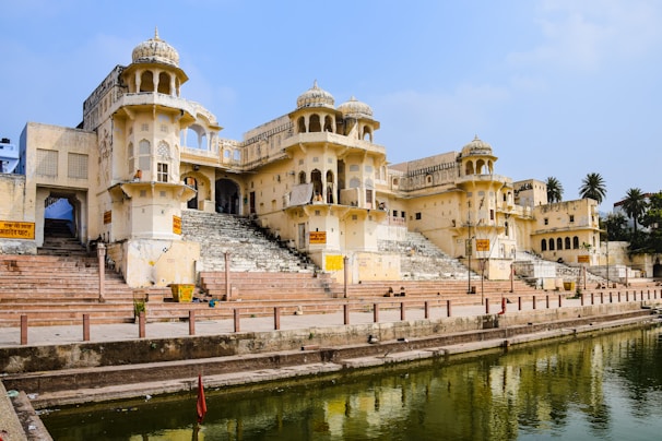 An ornate, historic building with intricate architectural details stands beside a serene, reflective body of water. The structure features multiple domes and arches typical of traditional Indian architecture, with a flight of wide stone steps leading down to the water. Palm trees and a clear blue sky are visible in the background, enhancing the picturesque setting.