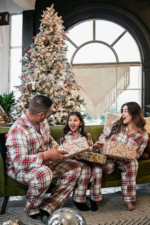 A family wearing matching plaid pajamas sits on a green couch in a festive living room. They are exchanging wrapped presents and appear joyful. Behind them is a decorated Christmas tree with red berries and white ornaments. A large window with a macram&eacute; wall hanging adds warmth to the scene.