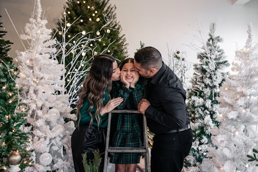 A family scene with two adults kissing a child's cheeks. The setting includes snowy Christmas trees adorned with lights, creating a festive and warm atmosphere. The family is gathered around a wooden ladder, with the child wearing a green plaid dress.