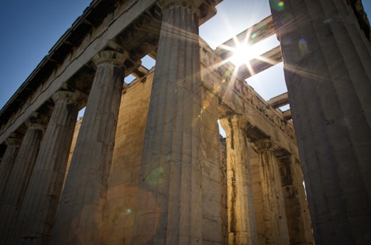 Ancient stone columns of a classical Greek temple with sunlight streaming between them, creating a dramatic lens flare effect. The architectural details of the columns and the clear blue sky add to the historical ambiance.