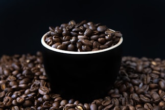 A black cup is filled with dark roasted coffee beans, sitting on a surface covered with more coffee beans. The background is a deep, dark color, enhancing the rich brown hues of the beans.