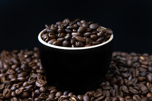 A black cup is filled with dark roasted coffee beans, sitting on a surface covered with more coffee beans. The background is a deep, dark color, enhancing the rich brown hues of the beans.