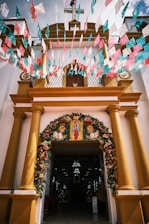 The entrance of a church is adorned with colorful floral decorations and papel picado banners in pink, blue, and white. The structure features yellow columns and a cross on top, with a religious image displayed above the entrance.