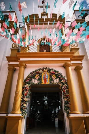 The entrance of a church is adorned with colorful floral decorations and papel picado banners in pink, blue, and white. The structure features yellow columns and a cross on top, with a religious image displayed above the entrance.