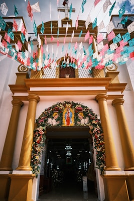 The entrance of a church is adorned with colorful floral decorations and papel picado banners in pink, blue, and white. The structure features yellow columns and a cross on top, with a religious image displayed above the entrance.