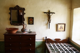 A modestly furnished room features a wooden dresser with a mirror above it, a ceramic jug, a bowl, a candle holder, and a crucifix mounted on the wall. A framed picture is on the wall next to the crucifix. A simple wooden bed frame is present without a mattress.