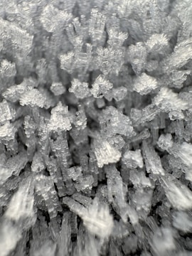 Close-up image of crystalline structures, possibly salt or sugar crystals, appearing in a chaotic and clustered arrangement. The crystals exhibit a transparent to opaque texture with sharp, angular edges.