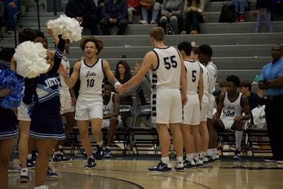 A basketball player wearing a 'Jets' uniform is running towards his teammates for a high five, while cheerleaders with pom-poms are performing nearby. Other players and coaching staff are seated on the bench, watching the ongoing activity. The scene is set in a gymnasium with spectators in the background.