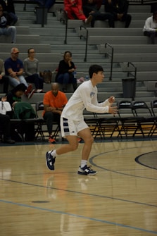 A basketball player in a white jersey and blue shoes is actively participating in a game. He appears focused and is mid-motion on the court. Behind him, spectators are seated on bleachers, watching the game intently.