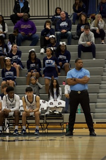 A basketball team is seated on the bench during a game. The players are dressed in uniform, accompanied by a coach standing beside them. Behind the bench, cheerleaders wearing blue uniforms with 'Jets' written on them are seated on bleachers. Spectators fill the bleachers, some wearing casual team-related attire.