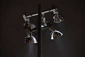A set of four metal floodlights mounted on a tall pole, casting beams of light in a diagonal direction against a dark night sky. The lamps have a shiny, reflective surface, and the scene conveys a sense of solitude or quiet.