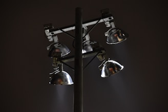 A set of four metal floodlights mounted on a tall pole, casting beams of light in a diagonal direction against a dark night sky. The lamps have a shiny, reflective surface, and the scene conveys a sense of solitude or quiet.