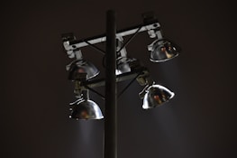 A set of four metal floodlights mounted on a tall pole, casting beams of light in a diagonal direction against a dark night sky. The lamps have a shiny, reflective surface, and the scene conveys a sense of solitude or quiet.