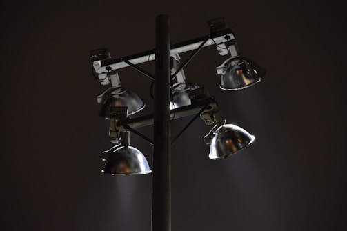 A set of four metal floodlights mounted on a tall pole, casting beams of light in a diagonal direction against a dark night sky. The lamps have a shiny, reflective surface, and the scene conveys a sense of solitude or quiet.