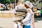 A street vendor carries a large stack of colorful hats on their arm. The vendor is wearing a hat similar to those they are selling and is dressed in a white shirt with a colorful scarf draped over the shoulder. The background shows a bustling outdoor setting with trees, people walking, and others sitting in a park-like area. Nearby, a backpack is visible hanging from a tree branch.
