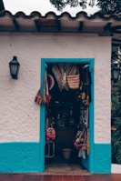 A small store with a white stucco facade and vibrant blue trim surrounds the doorway displaying various colorful woven bags and hats. The entrance is adorned with traditional Latin American crafts, suggesting a quaint, artisan shop vibe. The wall-mounted lanterns on either side hint at an inviting, rustic atmosphere.