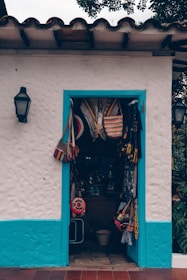 A small store with a white stucco facade and vibrant blue trim surrounds the doorway displaying various colorful woven bags and hats. The entrance is adorned with traditional Latin American crafts, suggesting a quaint, artisan shop vibe. The wall-mounted lanterns on either side hint at an inviting, rustic atmosphere.