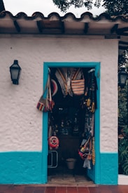 A small store with a white stucco facade and vibrant blue trim surrounds the doorway displaying various colorful woven bags and hats. The entrance is adorned with traditional Latin American crafts, suggesting a quaint, artisan shop vibe. The wall-mounted lanterns on either side hint at an inviting, rustic atmosphere.